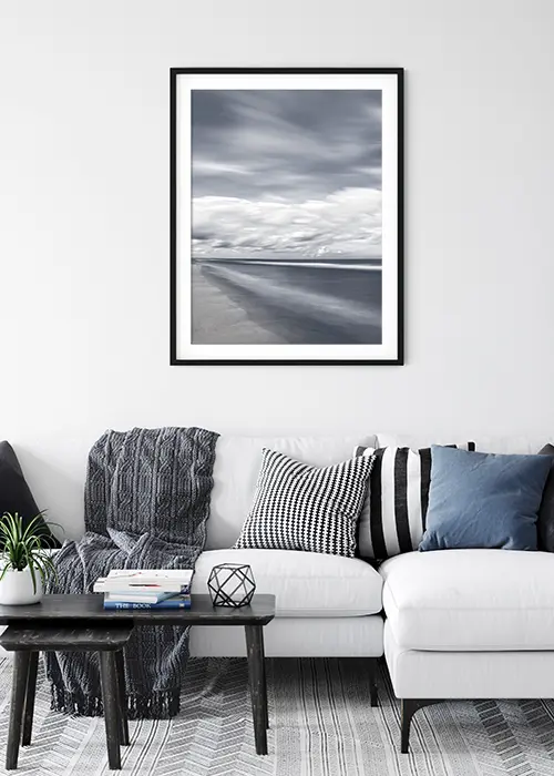 Poster mit Strand und Meer unter einem dramatischen Himmel in einer dezenten Blaustimmung über einem Sofa hängend