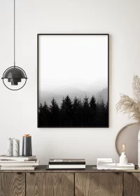Poster in schwarz-weiß mit einem Wald vor Bergen im Nebel über einem Sideboard hängend