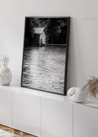 Poster mit einem alten Bootshaus an einem See in schwarz-weiß auf einem Sideboard an die Wand gelehnt