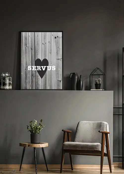 Poster mit einem schwarzen Herz und weißer Aufschrift Servus vor Holz-Hintergrund an eine graue Wand gelehnt