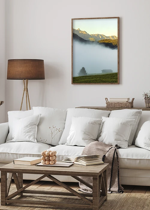 Poster mit einer im Nebel liegenden Alm und in der Sonne leuchtenden Berggipfeln über einem Sofa hängend